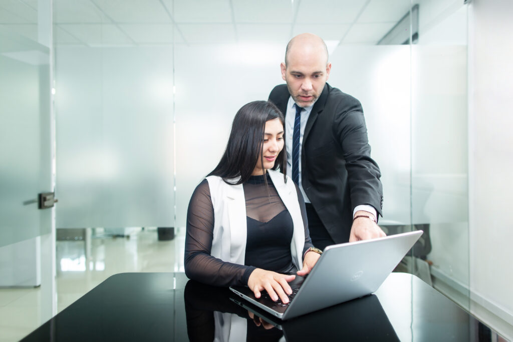 A man in a suit stands and points at a laptop screen while a woman, also in business attire, sits and types on the laptop. In their modern office with glass walls, both are focused on implementing payroll outsourcing in Guatemala.