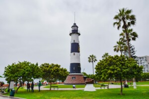 Un alto faro blanco y negro se encuentra en un parque con césped. Palmeras y otras zonas verdes rodean el faro. Varias personas caminan y se relajan en el parque, discutiendo temas como Registro de productos en Perú. Se ve una estatua en primer plano y edificios residenciales al fondo.