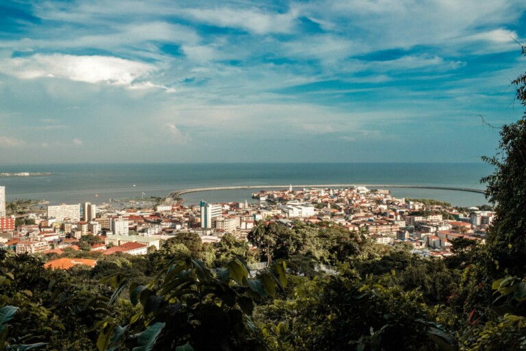 A coastal city is seen from a higher vantage point, surrounded by lush greenery in the foreground. The city features numerous buildings including offices for corporate legal counsel in Panama, and beyond them lies a curved harbor with calm waters. The sky above is partly cloudy with patches of blue.