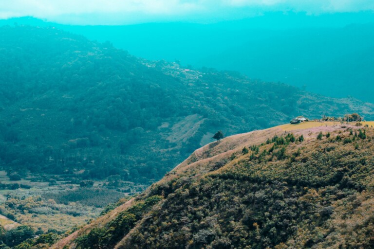 A scenic view of rolling green hills with a small structure perched near the top of one hill in Costa Rica. The foreground hill is covered in mixed vegetation, resembling the meticulous care seen in an Entity Health Check. The background is layered with additional hills under a hazy blue sky.