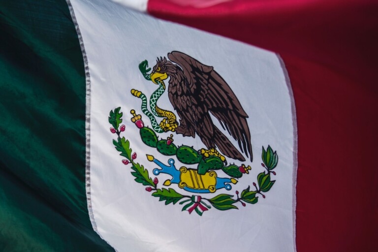 A close-up of the flag of Mexico. The flag features three vertical stripes: green on the left, white in the middle, and red on the right. In the center is the national emblem, depicting an eagle perched on a cactus with a snake in its beak and talon, symbolizing growth and strength to form a branch in Mexico.