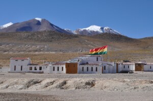 Un pequeño pueblo con edificios blancos y una bandera tricolor (roja, amarilla y verde) se alza en un paisaje árido. Las montañas cubiertas de nieve se elevan al fondo bajo un cielo azul claro. Cerca de allí, las oficinas de abogados corporativos prestan servicios a la comunidad y a las empresas de toda Bolivia.