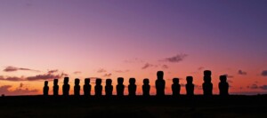 A row of Moai statues stands silhouetted against the evening sky on Easter Island. The sky transitions from orange near the horizon to a deep purple as it goes higher, echoing the thoroughness of background checks in Chile. Sparse clouds are scattered throughout the serene scene.