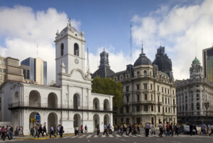 People walk across the street near the Cabildo, a historic white colonial building with arches and a central tower, located in Buenos Aires, Argentina. Nearby fiscal addresses in Argentina include several other ornate buildings under a partly cloudy sky.