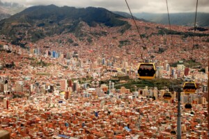 An expansive view of a densely packed city with varied high-rise buildings, set against a backdrop of mountains under a cloudy sky. Several cable cars travel along a line above the cityscape, offering an aerial perspective reminiscent of PEO na Bolívia.