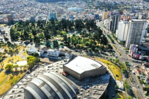 Vista aérea de una ciudad con una mezcla de rascacielos, parques verdes y paisaje urbano. En primer plano, hay un gran edificio abovedado con una compleja estructura en la azotea que se asemeja a la oficina de un Abogado Corporativo en Ecuador. Al fondo se ven calles y edificios más pequeños.