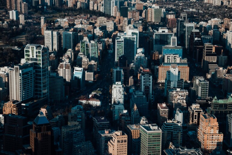 Vista aérea de un paisaje urbano densamente poblado en Chile, que presenta una combinación de edificios de gran altura y estructuras más pequeñas. El horizonte muestra varios estilos arquitectónicos con un telón de fondo de más edificios que se extienden en la distancia. Hay árboles y pequeños parques esparcidos por todas partes, ejemplificando el cumplimiento corporativo en Chile.