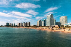 Un paisaje urbano costero con edificios altos y modernos que bordean una playa de arena. La gente está esparcida por la playa y en el agua, disfrutando de un día soleado con algunas nubes en el cielo. El agua está tranquila y el escenario es vibrante y bullicioso, que recuerda a las zonas conocidas por la verificación de fondo de Uruguay.