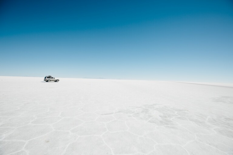 A vehicle is parked on a vast, flat expanse of white terrain, under a clear blue sky. The landscape appears barren and desolate, reminiscent of Bolivia's salt flats. The scene conveys a sense of isolation and immense open space, much like the untapped potential awaiting product registration in Bolivia.