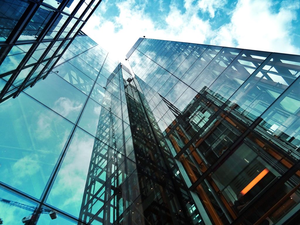 Uma fotografia tirada do chão para cima mostrando o exterior de vidro refletivo de vários prédios altos contra um céu azul com nuvens dispersas. As fachadas dos prédios se espelham umas nas outras, criando um padrão dinâmico de vidro e aço, muito parecido com um exame de saúde de uma entidade na paisagem urbana em expansão da Costa Rica.