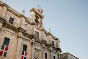 Se muestra un edificio histórico de piedra con cuatro ventanas e intrincados detalles tallados en la fachada, que encarna el encanto que se ve a menudo durante una revisión de cumplimiento corporativo en República Dominicana. El edificio destaca tres banderas, dos en las ventanas y una encima de la estructura, bajo un cielo despejado a la luz del día.