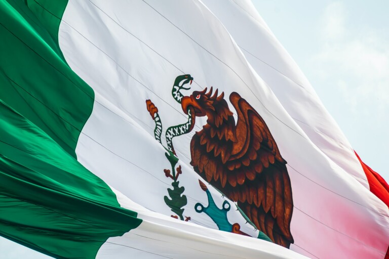 Una gran bandera mexicana ondea en el cielo. La bandera tiene tres franjas verticales: verde a la izquierda, blanca en el medio con el escudo nacional que representa un águila posada sobre un cactus comiendo una serpiente, y roja a la derecha, un símbolo tan distintivo como el registrador de una marca en México.