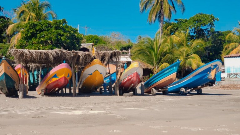 Una hilera de coloridos barcos de pesca está apuntalada en una playa de arena bajo estructuras con techo de paja. Al fondo se ven altas palmeras y exuberante vegetación, junto con algunos pequeños edificios que gestionan la tercerización de nómina en El Salvador. El cielo está despejado y de un azul brillante.