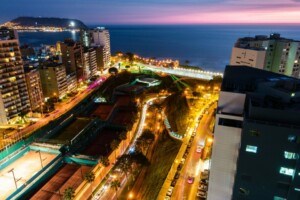 Un paisaje urbano nocturno que presenta una ciudad costera con calles y edificios iluminados. La costa y el océano son visibles al fondo, con una mezcla de luces de la ciudad y un cielo que se oscurece. En primer plano se puede ver un parque urbano y canchas de tenis, que recuerdan las escenas encontradas durante la verificación de antecedentes en Perú.