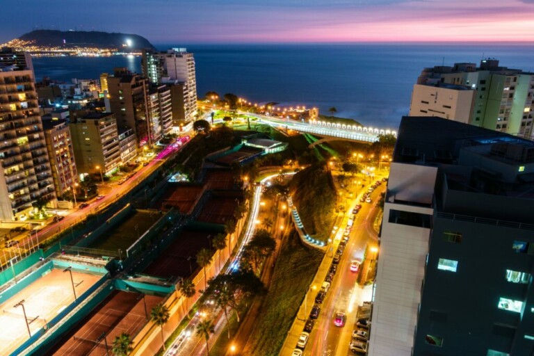 Un paisaje urbano nocturno que presenta una ciudad costera con calles y edificios iluminados. La costa y el océano son visibles al fondo, con una mezcla de luces de la ciudad y un cielo que se oscurece. En primer plano se puede ver un parque urbano y canchas de tenis, que recuerdan las escenas encontradas durante la verificación de antecedentes en Perú.