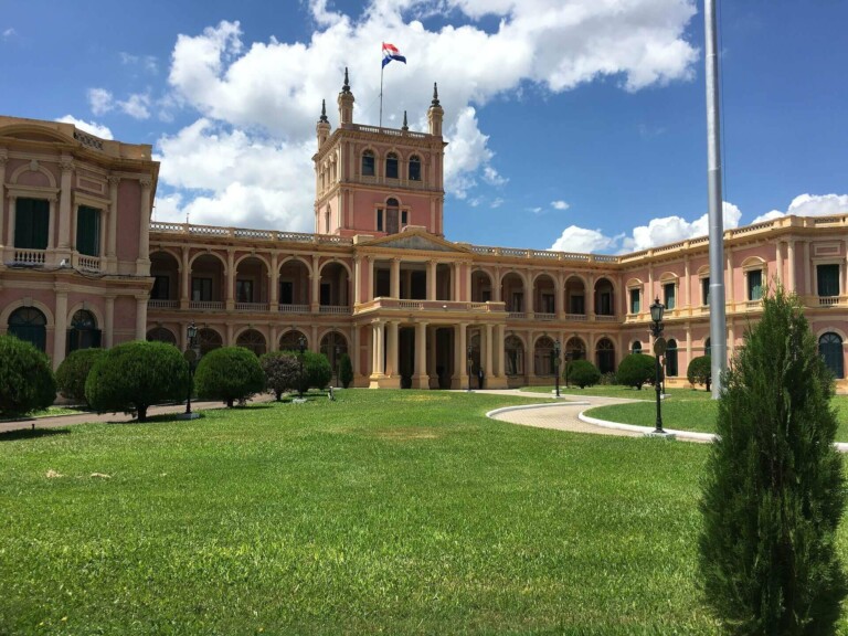 The image shows a large pink and beige historic building with arched windows, a central tower, and a flag on top. Surrounded by well-manicured lawns and bushes, the building creates an impressive backdrop for an empresa de folha de pagamento e PEO no Paraguai. A flagpole is also visible in the foreground beneath a partly cloudy sky.