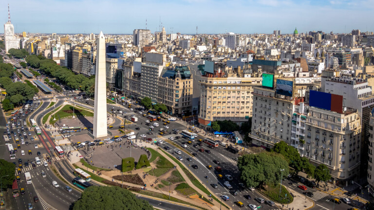 Vista aérea de la bulliciosa ciudad de Buenos Aires con el Obelisco de Buenos Aires en el centro de una plaza circular. Alrededor de la plaza hay calles muy transitadas con numerosos vehículos. La zona está densamente poblada de edificios e infraestructura urbana.