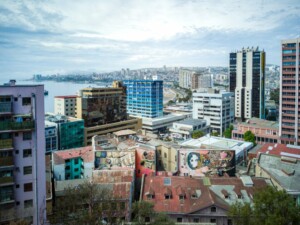 Una vista del paisaje urbano de Valparaíso, Chile, que muestra una mezcla de edificios modernos y antiguos con coloridos murales. En el horizonte predominan diversos estilos arquitectónicos, con la costa al fondo bajo un cielo parcialmente nublado. Son escenas como esta las que atraen a muchos a comprar una propiedad en Santiago.