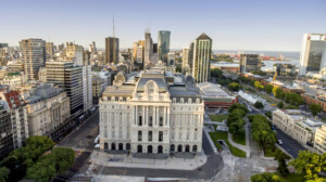 Aerial view of a large, historic building with intricate architectural details set amid a bustling cityscape. Surrounding the building are modern high-rises, green parks with trees, and a waterfront area featuring ships and cranes in the background—an ideal setting for company incorporation in Argentina.