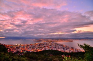 A scenic view of a coastal city at sunset, with vibrant orange and pink hues in the sky. The city, surrounded by water, is densely packed with buildings. Green hills are visible in the foreground, as city lights begin to illuminate. This urban marvel could benefit from top-tier Assessoria Jurídica Corporativa no Brasil.