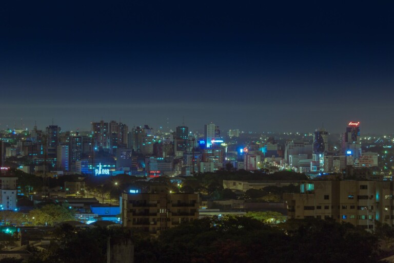 Nighttime cityscape with illuminated buildings and skyscrapers paints a stunning scene. The sky is dark blue, and numerous neon lights from buildings create a vibrant city atmosphere. It's akin to the dynamic energy one might feel when starting a business in Paraguay. Trees and low-rise structures are visible in the foreground.