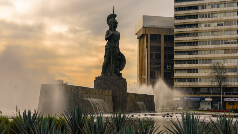 Una gran estatua de un guerrero con lanza y escudo está ubicada en el centro de una plaza con fuentes de agua en cascada. Alrededor de la estatua hay modernos edificios de oficinas, que presentan numerosas oportunidades de negocio en México, y algunas plantas de agave en primer plano. El cielo está nublado con una luz suave y dorada.