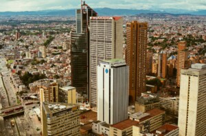 Vista de un paisaje urbano con varios edificios altos, incluido un prominente rascacielos negro y varias otras estructuras de gran altura. El área circundante está densamente poblada de más edificios, y las montañas son visibles a lo lejos bajo un cielo nublado, perfecto para aquellos que buscan una visa de negocios en Colombia.