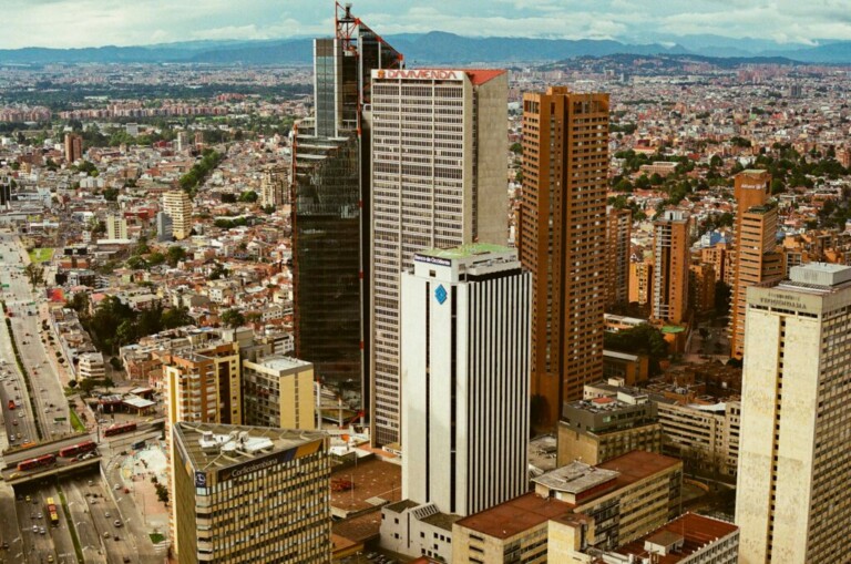 Vista de un paisaje urbano con varios edificios altos, incluido un prominente rascacielos negro y varias otras estructuras de gran altura. El área circundante está densamente poblada de más edificios, y las montañas son visibles a lo lejos bajo un cielo nublado, perfecto para aquellos que buscan una visa de negocios en Colombia.