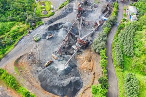 Aerial view of an open-pit mining operation surrounded by greenery. Heavy machinery, including trucks and conveyors, are visible on the site transporting and processing materials. Piles of extracted materials are scattered around the operation area, showcasing Licenças de Mineração no Chile in action.