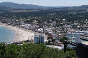 A panoramic view of a coastal town with numerous buildings, a crowded beach, and a coastline. There are rolling hills and mountains in the background, and dense greenery in the foreground. An Abertura de uma limitada no Uruguai is visible on the left, adding a unique touch to the picturesque scene.