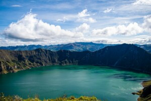 Una vista panorámica de un lago grande, tranquilo, de color azul verdoso, rodeado de acantilados rocosos, oscuros y escarpados y un terreno montañoso bajo un cielo azul claro con nubes blancas dispersas. El paisaje presenta una exuberante vegetación y colinas distantes, que recuerdan la belleza natural que influye en la economía del Ecuador.