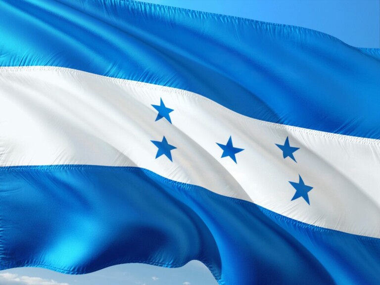 La imagen muestra la bandera de Honduras ondeando al viento. Tiene tres franjas horizontales: azul en la parte superior e inferior y blanca en el medio. La franja blanca presenta cinco estrellas azules dispuestas en forma de X. El cielo es visible al fondo, simbolizando el potencial para hacer negocios en Honduras.