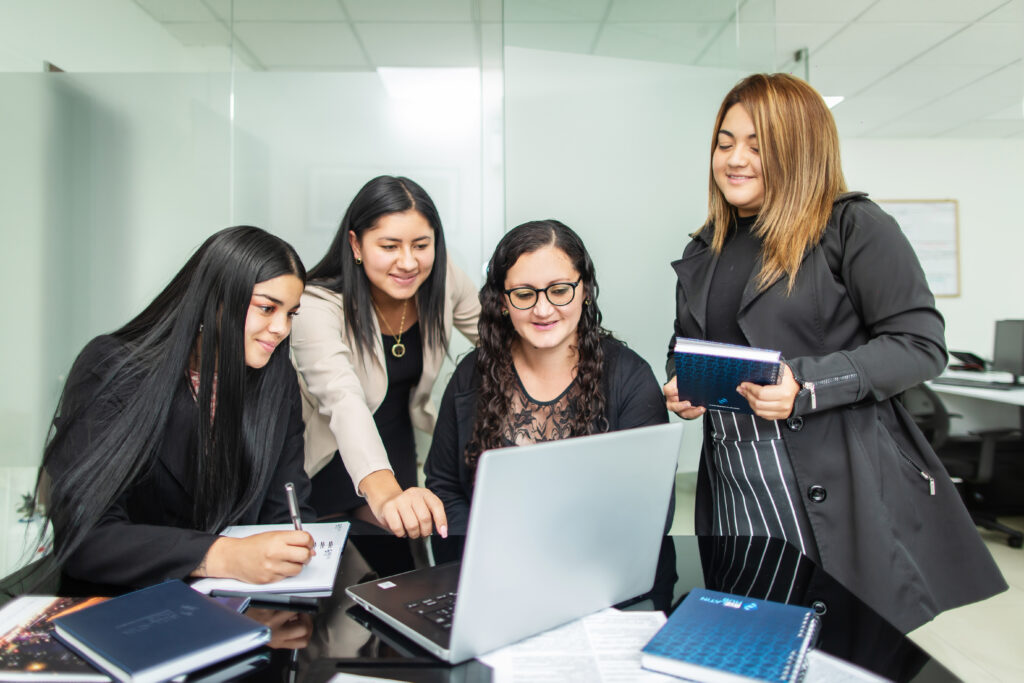 Four women are gathered around a table in an office setting at an accounting firm in Chile, interacting with a laptop. One woman is pointing at the screen while two others look on. The fourth woman holds a notebook and smiles. There are notebooks and papers on the table.