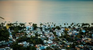 Un pueblo costero de México con una colección de coloridos edificios y casas se ve desde un mirador elevado. Las palmeras se alinean en la orilla y, al fondo, se ven pequeños barcos esparcidos por las tranquilas aguas. El cielo y el agua reflejan tonos de rosa y azul.