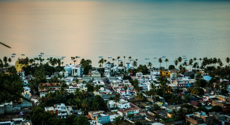 Un pueblo costero de México con una colección de coloridos edificios y casas se ve desde un mirador elevado. Las palmeras se alinean en la orilla y, al fondo, se ven pequeños barcos esparcidos por las tranquilas aguas. El cielo y el agua reflejan tonos de rosa y azul.