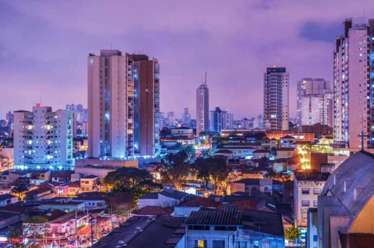 Un paisaje urbano al anochecer con numerosos edificios altos, ventanas iluminadas y farolas vibrantes. En primer plano se ven casas residenciales y árboles verdes, mientras que rascacielos más grandes ocupan el fondo bajo un cielo de tonos violetas, encarnando el espíritu de Debida Diligencia en Brasil.