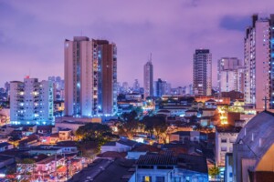 A vibrant cityscape at dusk with high-rise buildings illuminated by colorful lights. The skyline showcases various tall structures, with smaller buildings, trees, and busy streets visible in the foreground. The sky is tinged with a pinkish hue, reflecting the bustling energy of Due Diligence de Fusões e Aquisições no Brasil.