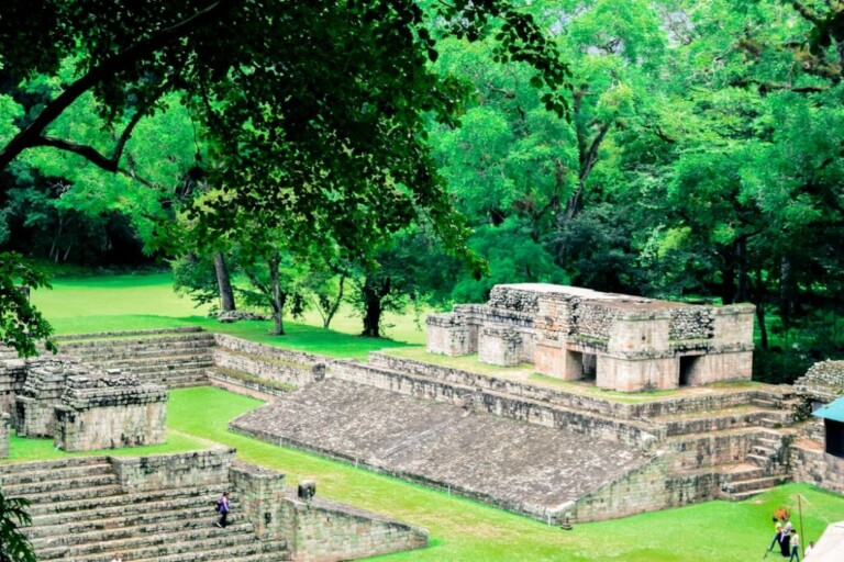 Antiguas ruinas de piedra, rodeadas de exuberante vegetación, cuentan con niveles de plataformas y escalones que conducen a una estructura central. Se puede ver a varias personas explorando el sitio, destacando su importancia histórica y su entorno natural: inspiración perfecta para quienes buscan crear una empresa en Honduras. Los árboles enmarcan el primer plano.