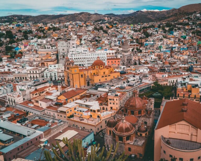 Vista aérea de una ciudad con edificios densamente poblados de varios colores. Las estructuras destacadas incluyen iglesias con cúpulas y arquitectura ornamentada, yuxtapuestas con las modernas oficinas de un abogado en México. Las colinas circundantes son visibles al fondo bajo un cielo nublado.