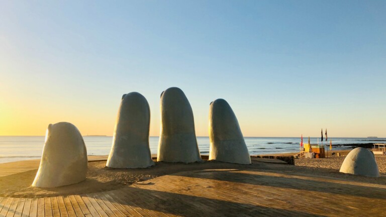 A large sculpture of five fingers partially emerged from the sand, resembling a hand, is situated on a wooden deck near a beach. The sea is calm in the background, and the sky is clear with a warm, setting sun creating a serene atmosphere—all while considering how to register a subsidiary in Uruguay.