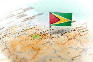 Una pequeña bandera de Guyana se coloca en un mapa, indicando su ubicación en América del Sur. El mapa muestra países vecinos, incluido Surinam, y regiones como las Tierras Altas de Guayana. Varios detalles geográficos, ciudades y áreas ricas en petróleo en Guyana también son visibles alrededor del área.