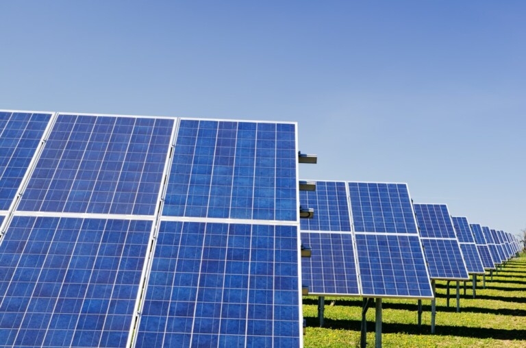 Una fila de paneles solares está colocada en un campo, con un cielo azul claro en lo alto. Los paneles están inclinados hacia el sol, capturando la energía solar. El suelo está cubierto de hierba verde, lo que crea una atmósfera luminosa y soleada: una inversión verde ejemplar.