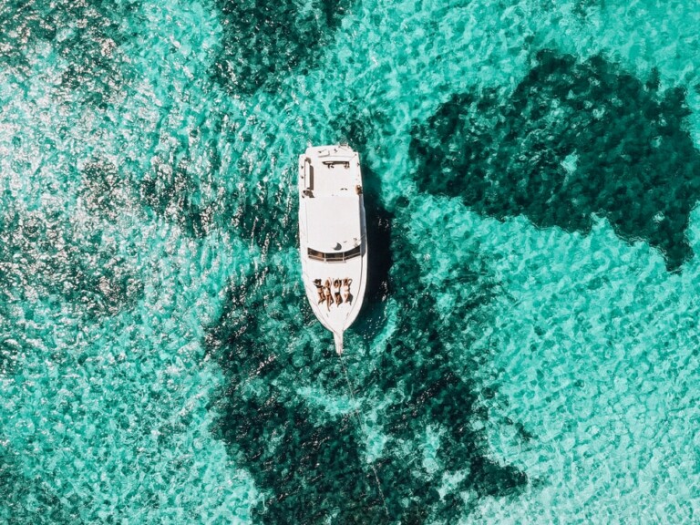 Vista aérea de un barco blanco con gente a la deriva en aguas cristalinas de color turquesa cerca de PEO en Honduras. La luz del sol se refleja en el agua, creando un efecto brillante. Las manchas oscuras en el agua sugieren diferentes profundidades o formaciones submarinas.