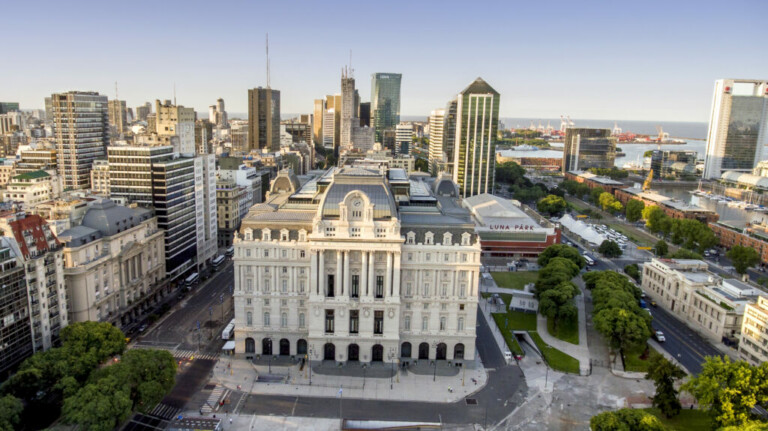 Una vista aérea de un gran edificio histórico rodeado de modernos edificios de gran altura en un paisaje urbano. El edificio, que recuerda a los primeros días cuando florecía la Constitución de empresas en Argentina, presenta una arquitectura clásica con columnas y una cúpula central. A la derecha se ven árboles y un área de parque, y al fondo hay una masa de agua.