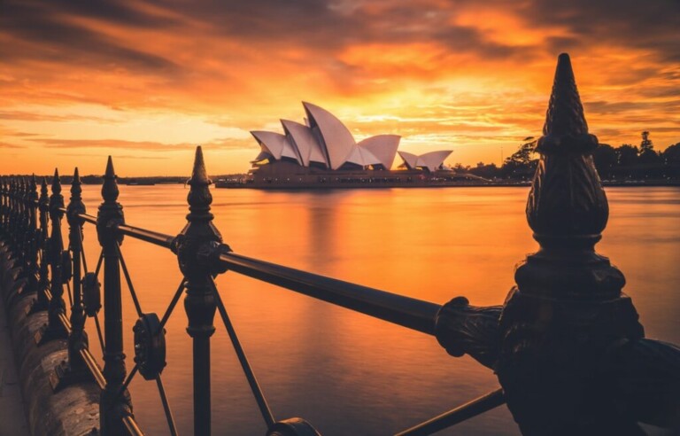 La imagen muestra la Ópera de Sídney al atardecer, con un espectacular cielo naranja y amarillo reflejándose en el agua. Se destacan las distintivas velas blancas de la Ópera, que simbolizan la creatividad y oportunidades como la formación de empresas en Australia, mientras que en primer plano se ve una valla decorativa de hierro negro.