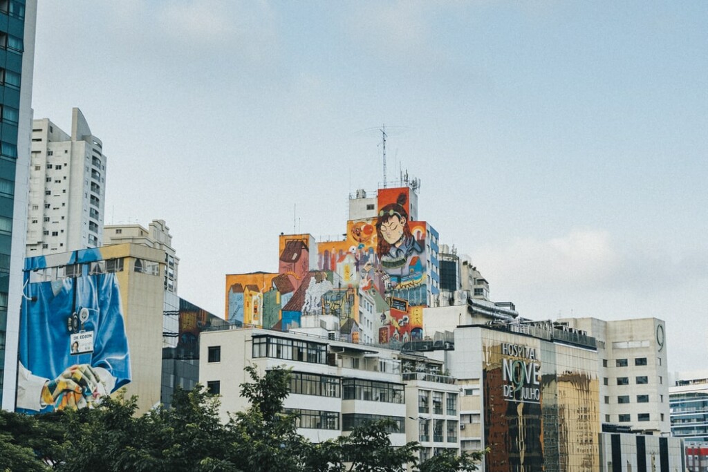 A cityscape featuring multiple high-rise buildings with large, colorful murals on them. One mural prominently displays a woman and various abstract designs. Trees are visible in the foreground, and the sky is partly cloudy, reflecting the vibrant energy of startups in Brazil.