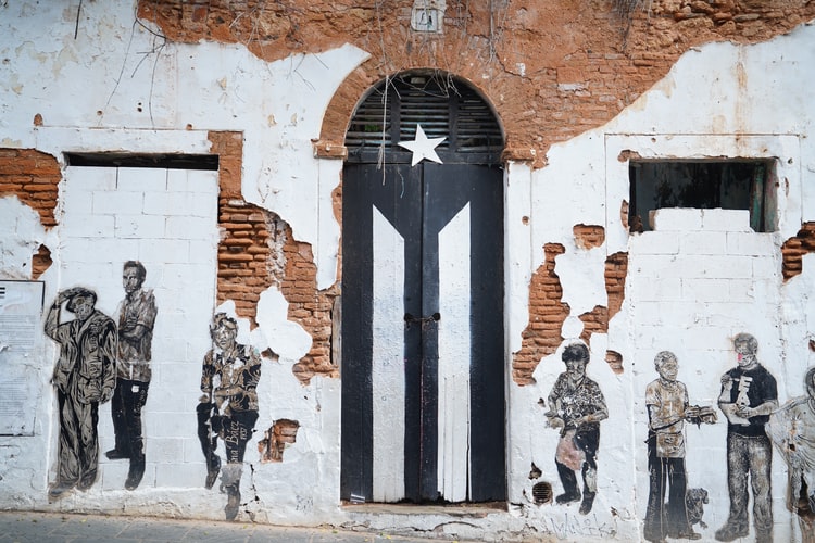 Una pared desgastada presenta figuras de personas pintadas en blanco y negro a ambos lados de una puerta que está pintada para parecerse a la bandera puertorriqueña, lo que refleja el espíritu de PEO en Puerto Rico. La pared muestra signos de deterioro con ladrillos a la vista y yeso desmoronado.