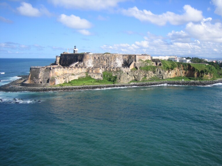 Un gran fuerte de piedra con un faro se alza sobre un acantilado cubierto de hierba con vista al océano, ofreciendo *oportunidades de negocio* únicas en Puerto Rico. El cielo está parcialmente nublado y las olas rompen contra la orilla. Los edificios se ven apenas visibles al fondo.