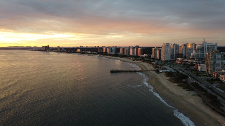 Aerial view of a coastal city during sunset, showing high-rise buildings near the shoreline with a long pier extending into the water. The sky is partly cloudy, and waves gently reach the sandy beach. The city's skyline, reminiscent of places where Verificações de antecedentes no Uruguai might take place, is visible in the distance.