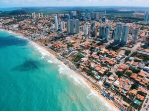 Aerial view of a coastal city in Brasil with numerous tall buildings and residential areas next to a sandy beach. The beach is lined with umbrellas, and the turquoise ocean has visible waves. The city extends inland, surrounded by vegetation and water bodies, reflecting the confianza de inversores.